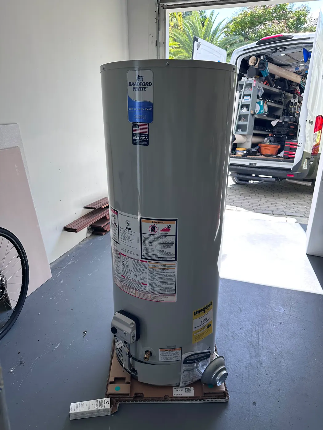 A new gas water heater standing in a garage with the open The God Plumber work van behind it