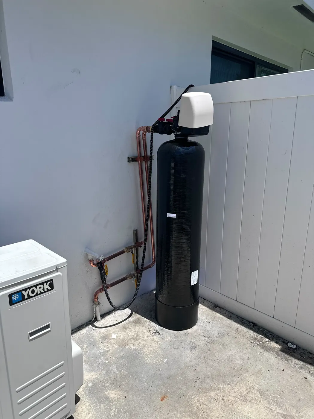 Black whole-house water softener tank installed outside next to an air-conditioning unit, with new copper connections